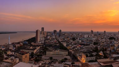 A cityscape at sunset with a river in the foreground and a mix of high-rise and low-rise buildings.