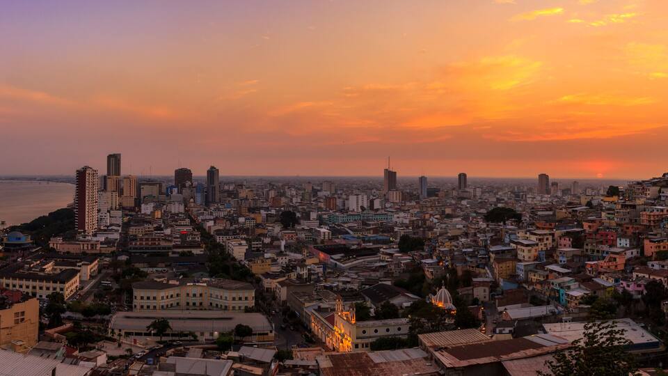 A cityscape at sunset with a river in the foreground and a mix of high-rise and low-rise buildings.