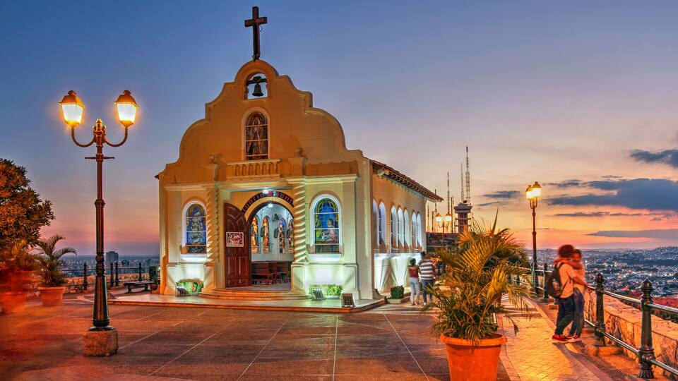 A church with a cross, arched windows, and a bell tower, surrounded by potted plants and street lamps.