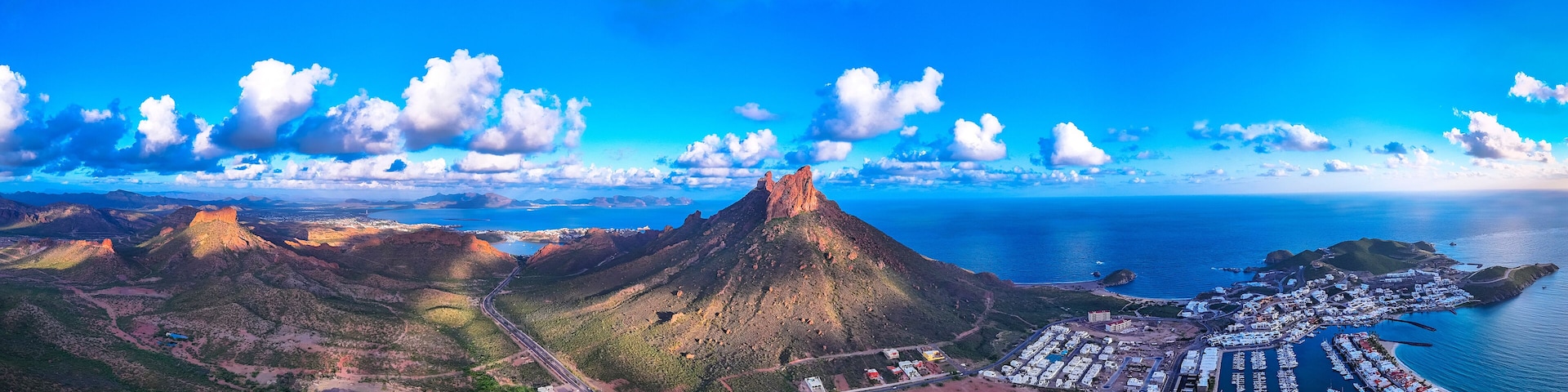 Un pueblo costero con una montaña imponente, un puerto deportivo y una carretera sinuosa.