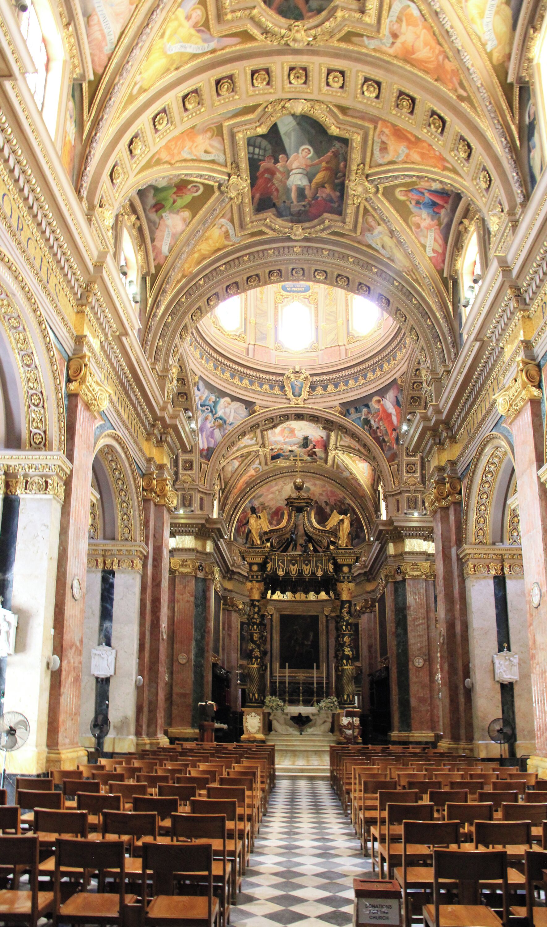 An ornate church interior with a high ceiling, detailed frescoes, and rows of wooden pews.