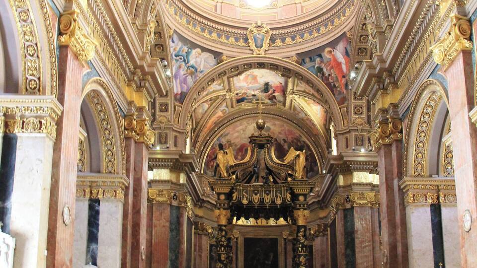 An ornate church interior with a high ceiling, detailed frescoes, and rows of wooden pews.