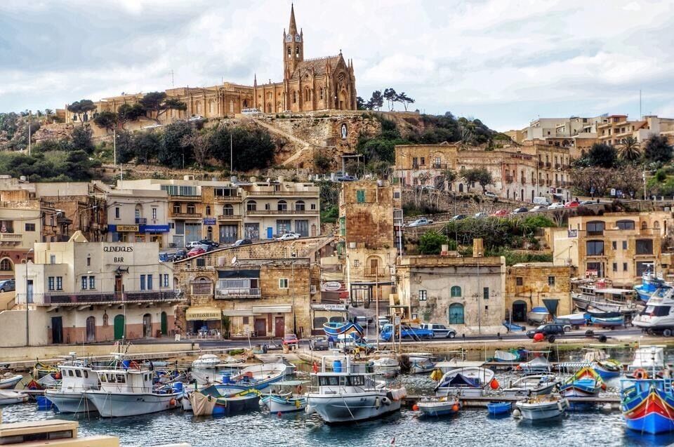 A coastal town with historic buildings, a prominent church on a hill, and a marina with boats.