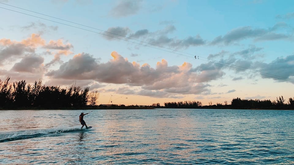 A person wakeboarding on a lake with a sunset sky and trees in the background.