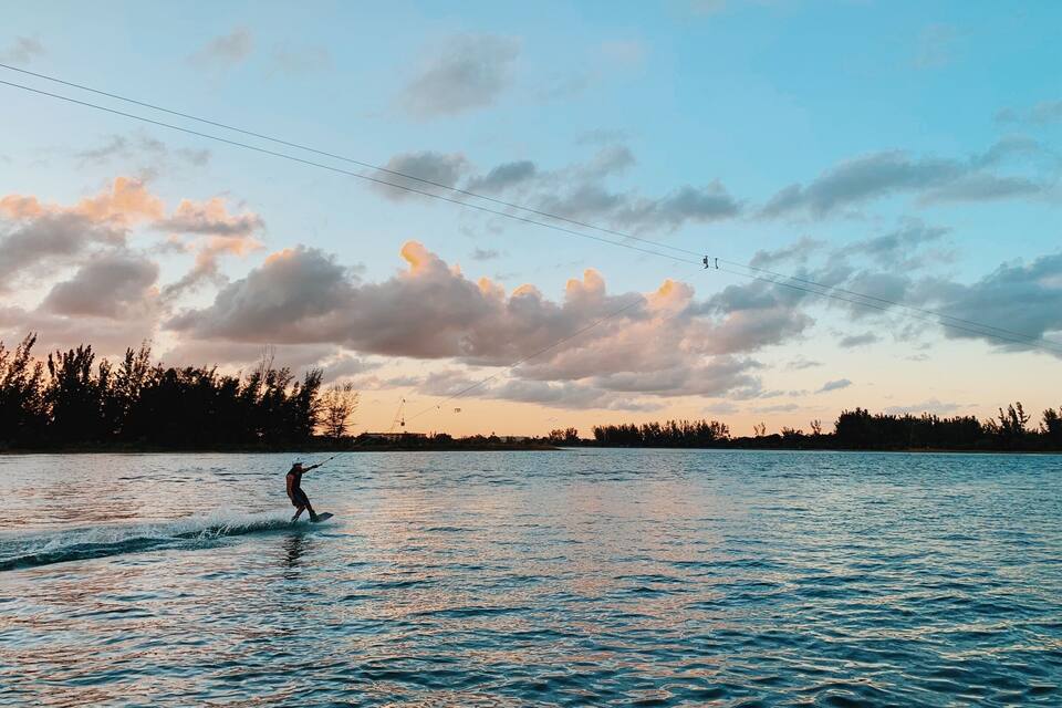 Iemand wakeboard op een meer, met een zonsondergang aan de hemel en bomen op de achtergrond.
