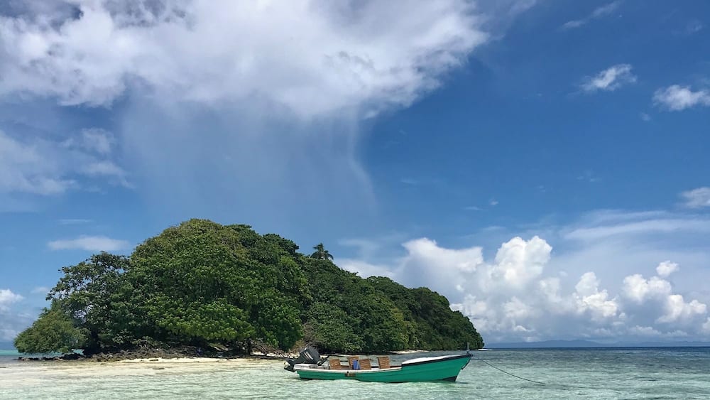A small island with dense green foliage, a boat anchored near the shore, and clear turquoise waters.