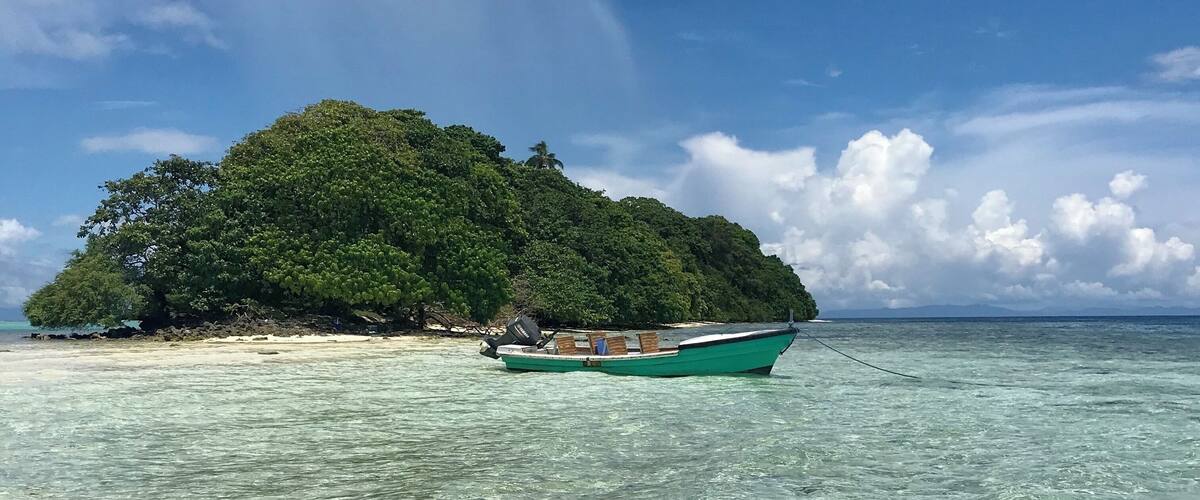 A small island with dense green foliage, a boat anchored near the shore, and clear turquoise waters.