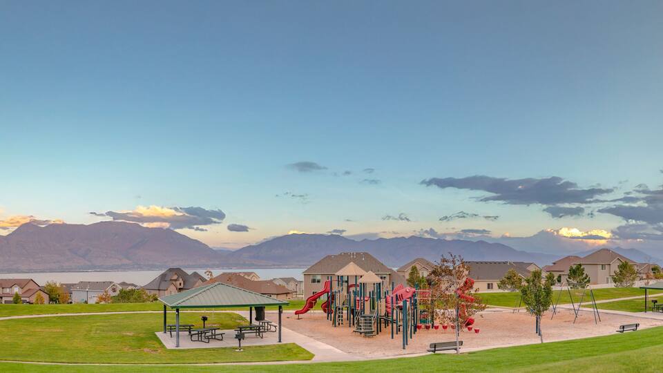 A playground with slides and swings, a picnic area, and houses in the background with mountains and a lake.