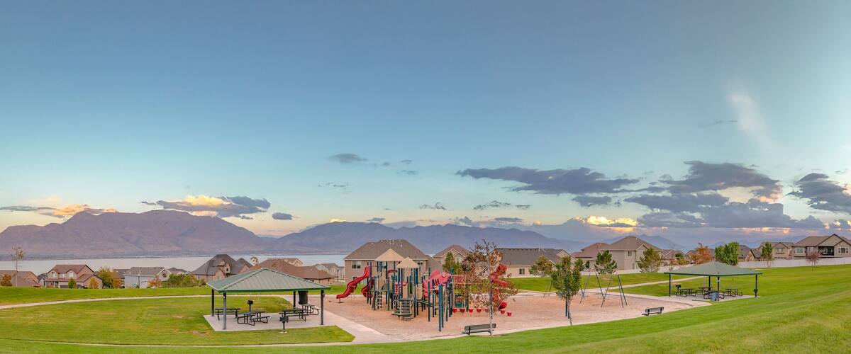 A playground with slides and swings, a picnic area, and houses in the background with mountains and a lake.