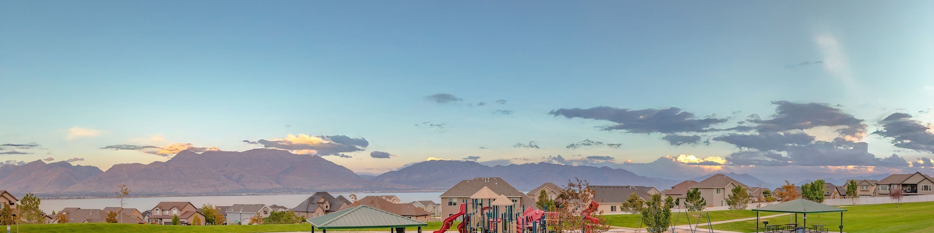 A playground with slides and swings, a picnic area, and houses in the background with mountains and a lake.