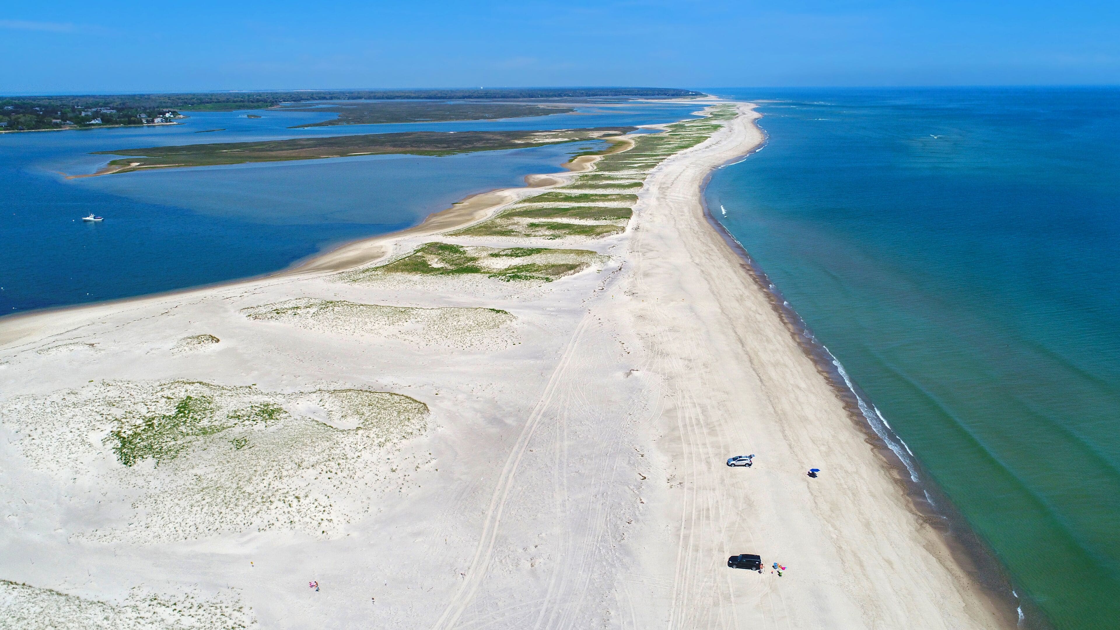 A coastal landscape with a long sandy beach, clear blue waters, and a few vehicles on the shore.