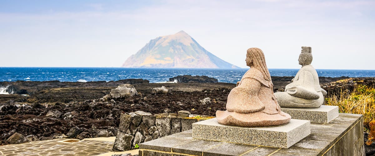 Two stone statues on a stone platform overlooking a rocky coastline with a mountain in the background.
