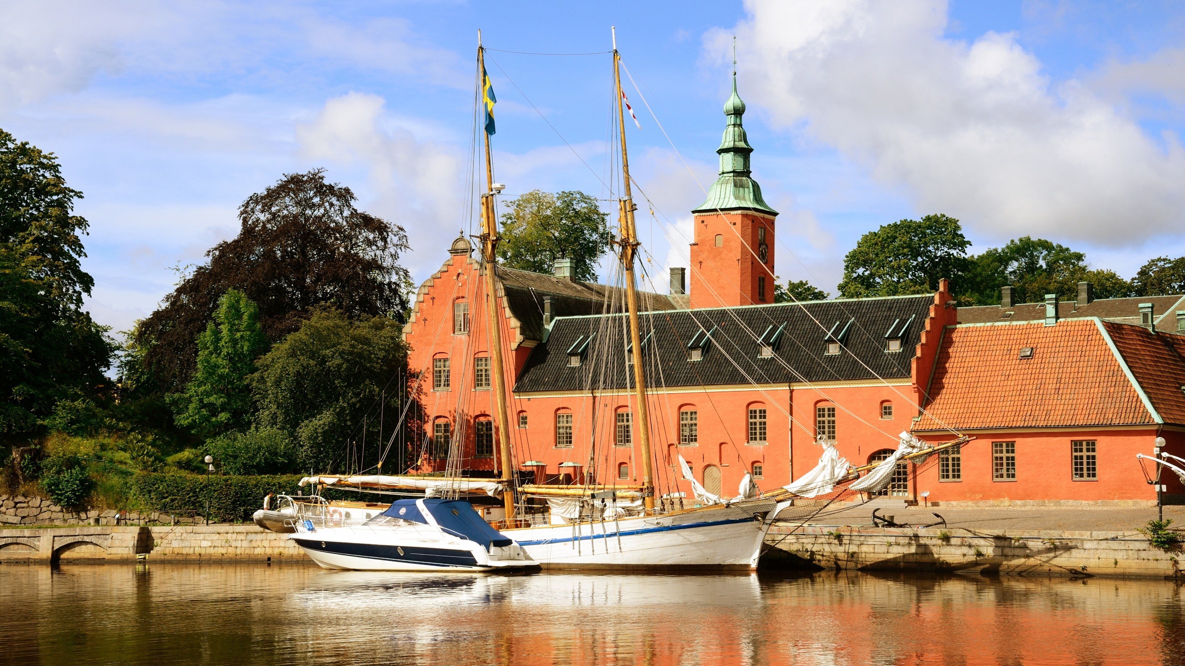 A historic building with a red facade and a green spire, a traditional boat docked in front, and a stone bridge in the background.