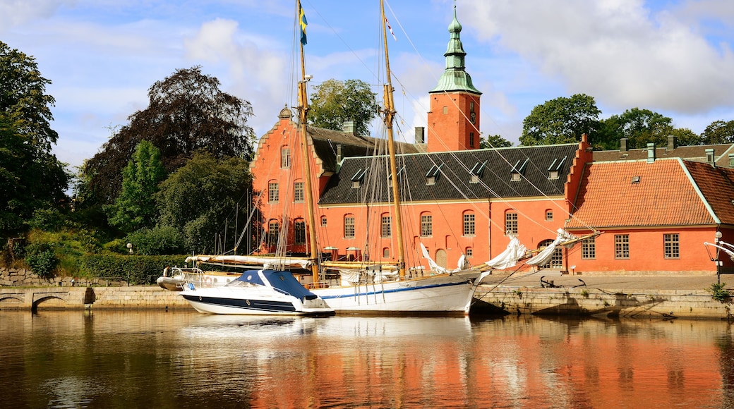 Edifício histórico com fachada vermelha e torre verde, um barco branco atracado em frente e um rio calmo refletindo a paisagem.