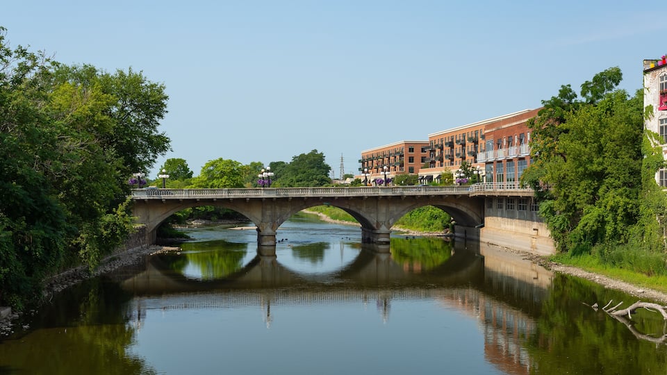 A stone bridge over a calm river, surrounded by lush greenery and buildings.