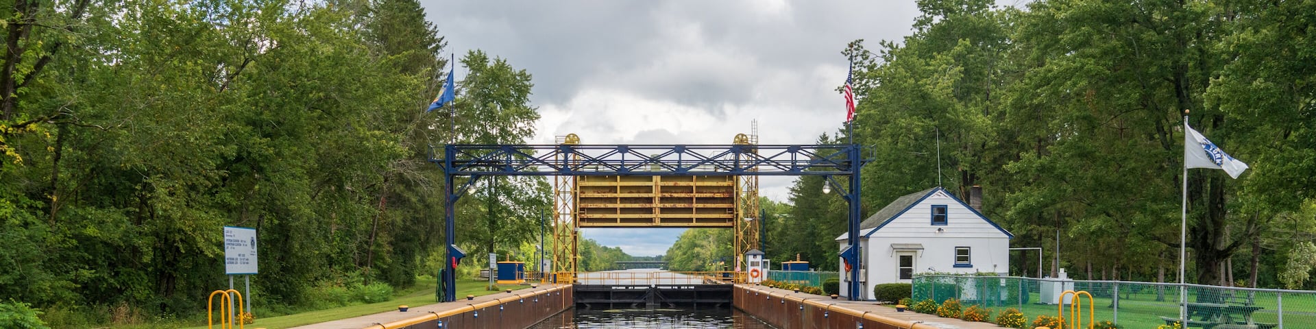A lock gate in a canal with a bridge overhead, surrounded by greenery and a small white building.