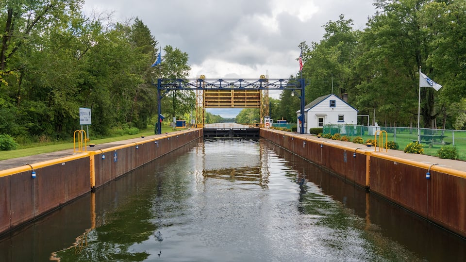 A lock gate in a canal with a bridge overhead, surrounded by greenery and a small white building.