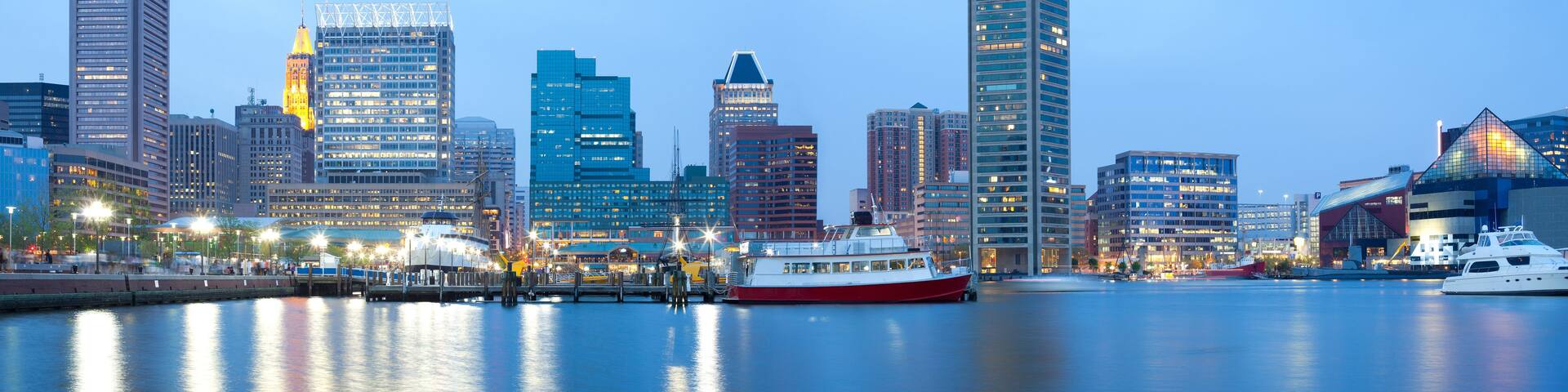 A waterfront cityscape with modern buildings, a red and white boat docked at the pier, and a clear sky.