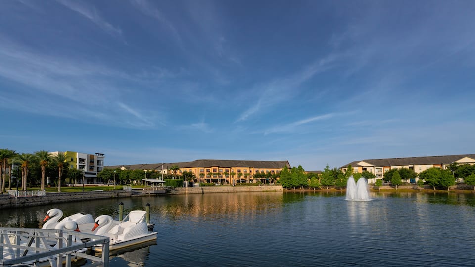A calm lake with swans on a dock, a fountain, and a building complex in the background.