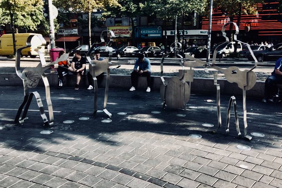A public square with modern art sculptures, a busy street with cars, and shops in the background.