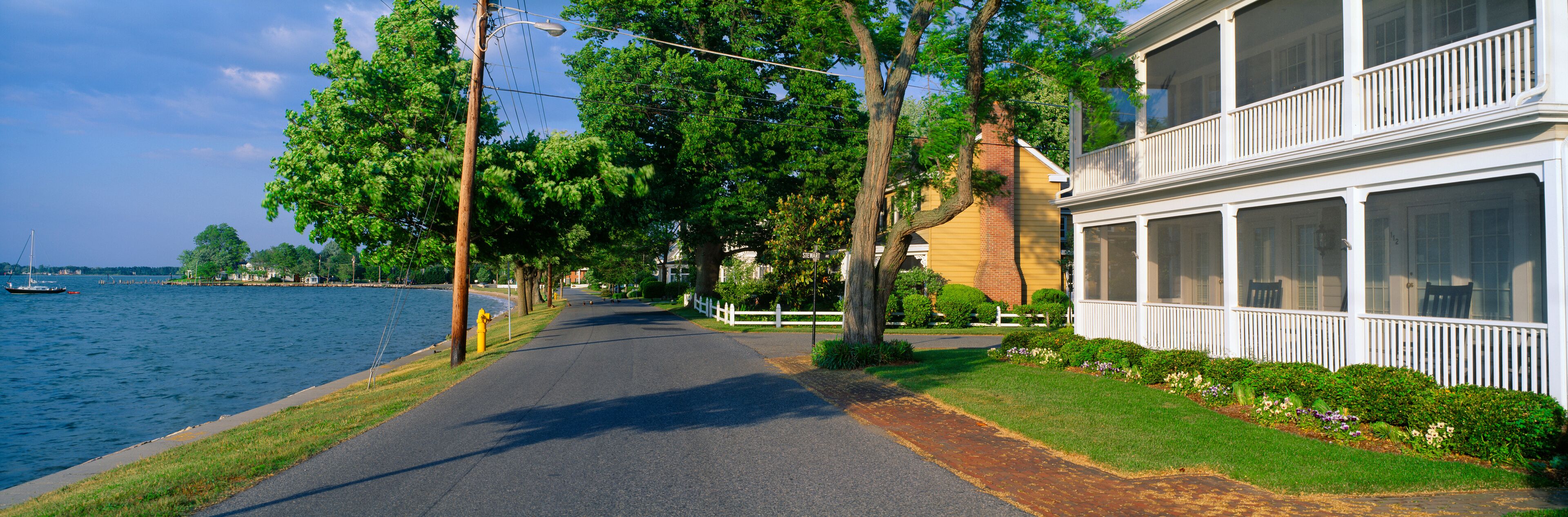 A waterfront street lined with trees and a white building with a balcony.