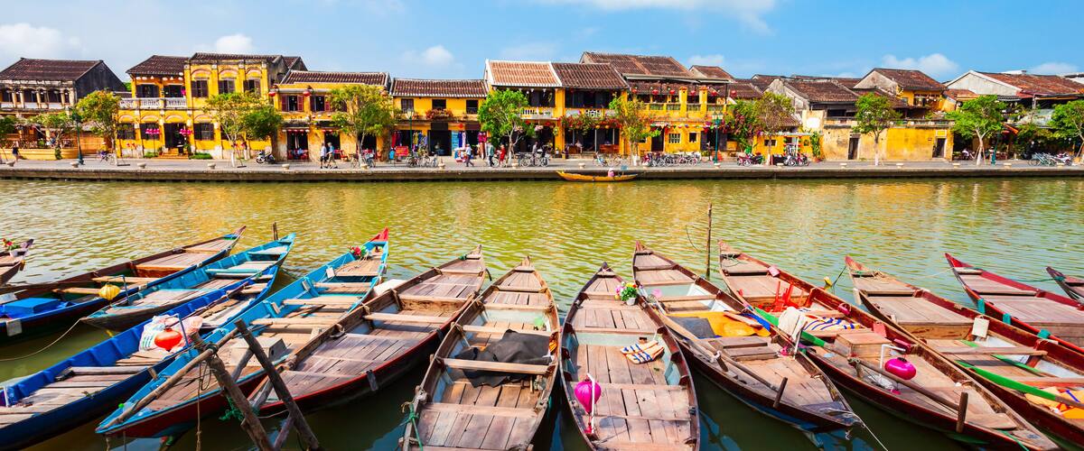 A row of colorful wooden boats docked along a riverbank with traditional yellow buildings in the background.