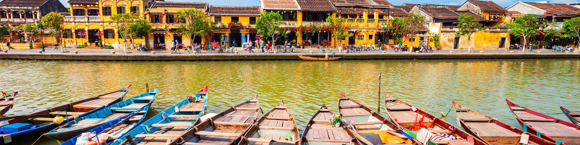 A row of colorful wooden boats docked along a riverbank with traditional yellow buildings in the background.