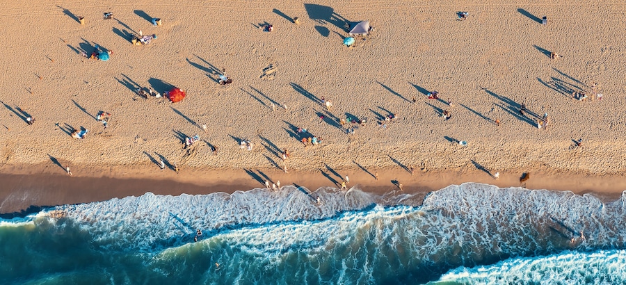Eine Vogelperspektive zeigt einen Strand mit Menschen, Sonnenschirmen und Wellen.