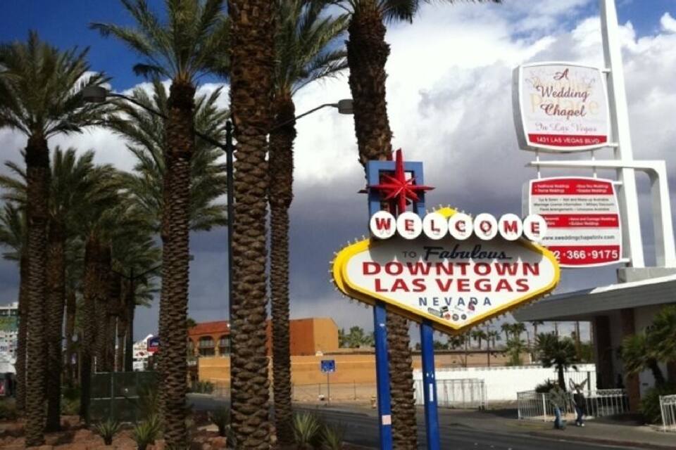 Placa de 'Bem-vindos ao centro de Las Vegas' com palmeiras e uma igreja ao fundo.
