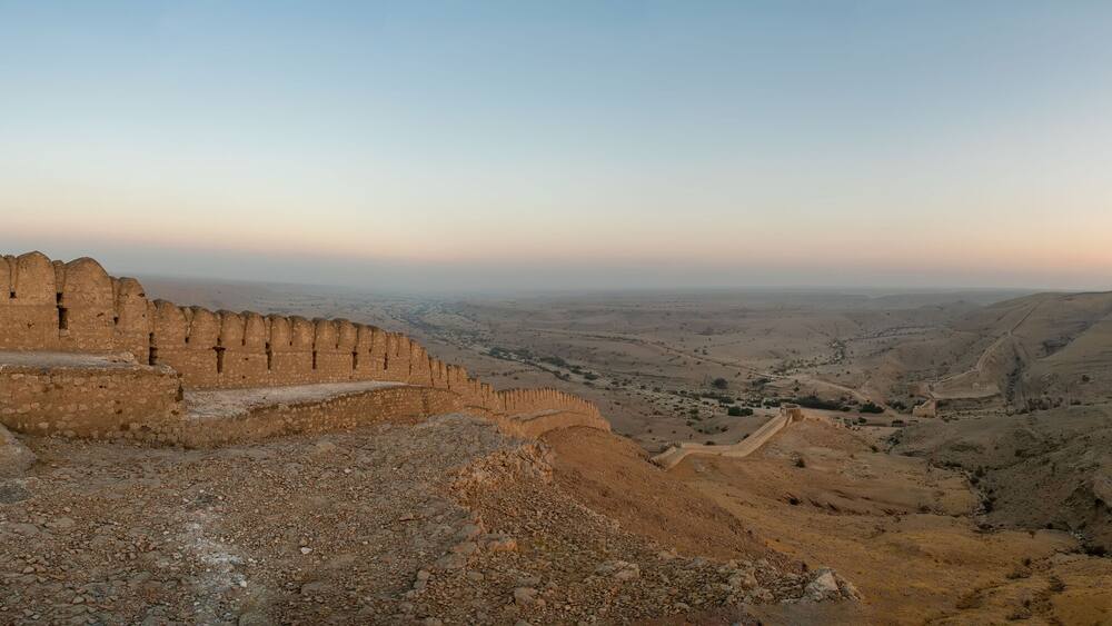 A rugged landscape with a prominent fortification on the left and a hilltop structure in the distance.