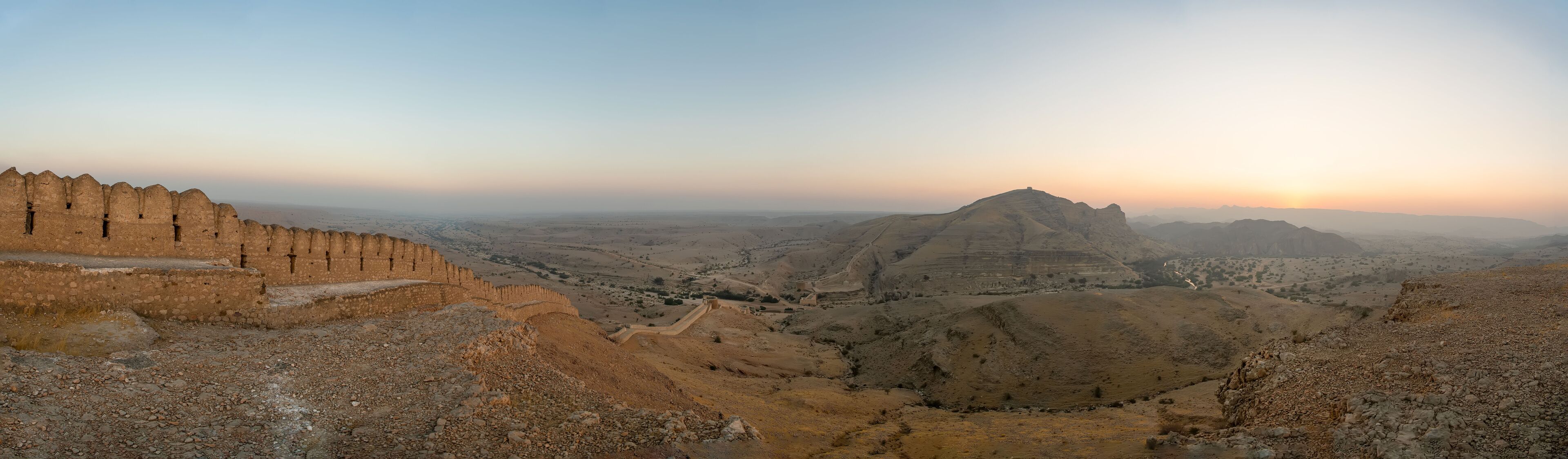 A rugged landscape with a prominent fortification on the left and a hilltop structure in the distance.