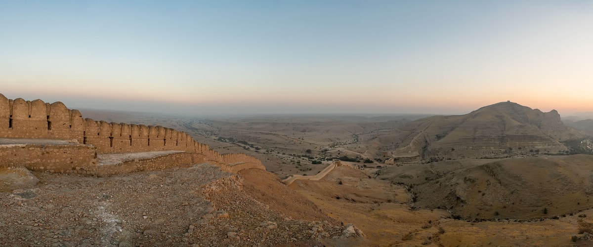 A rugged landscape with a prominent fortification on the left and a hilltop structure in the distance.