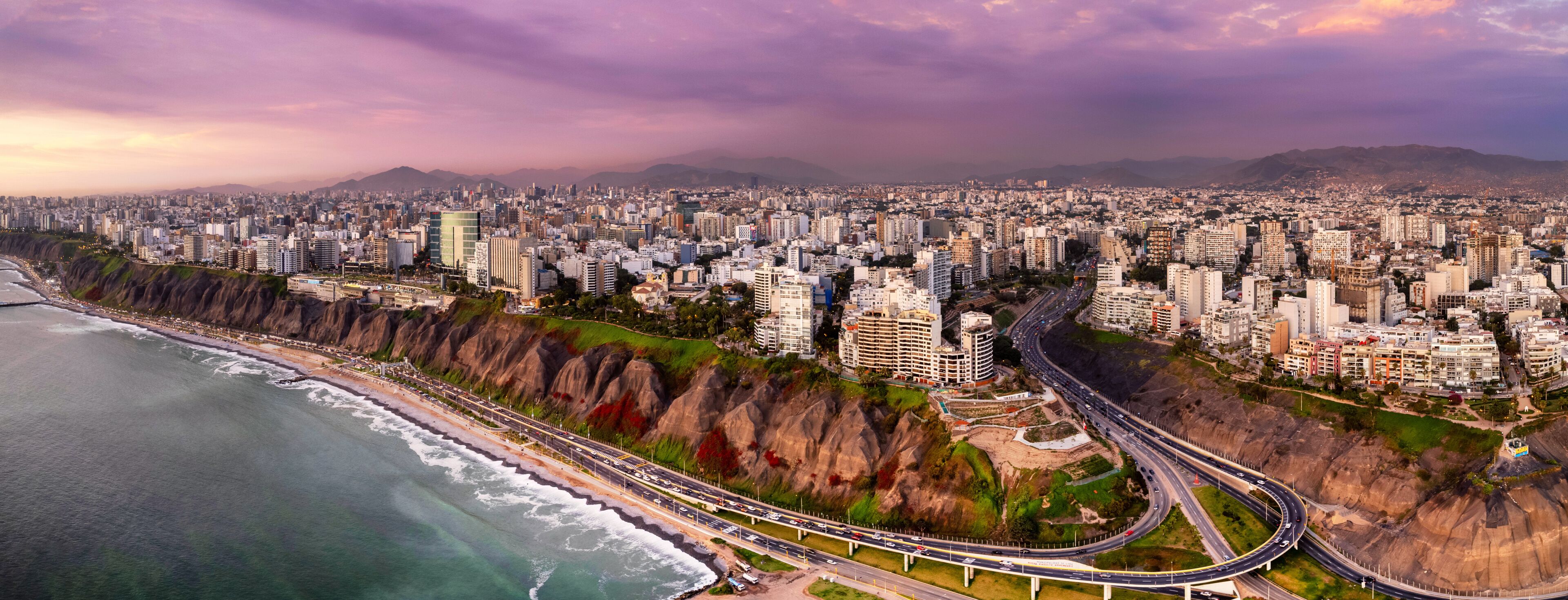A coastal cityscape with a prominent highway curving along the coastline, dense urban buildings, and a mountain range in the distance.