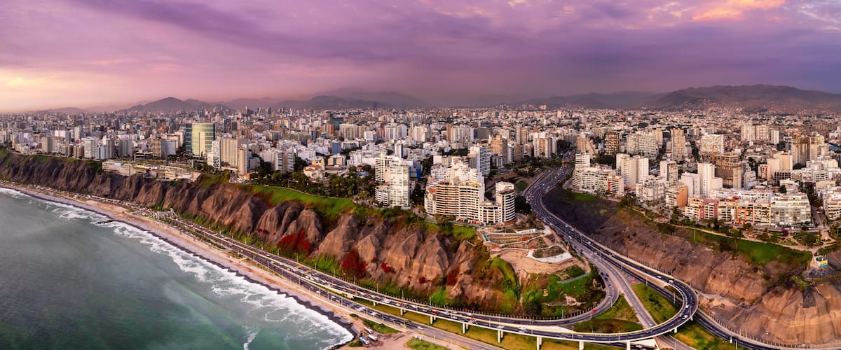 A coastal cityscape with a prominent highway curving along the coastline, dense urban buildings, and a mountain range in the distance.