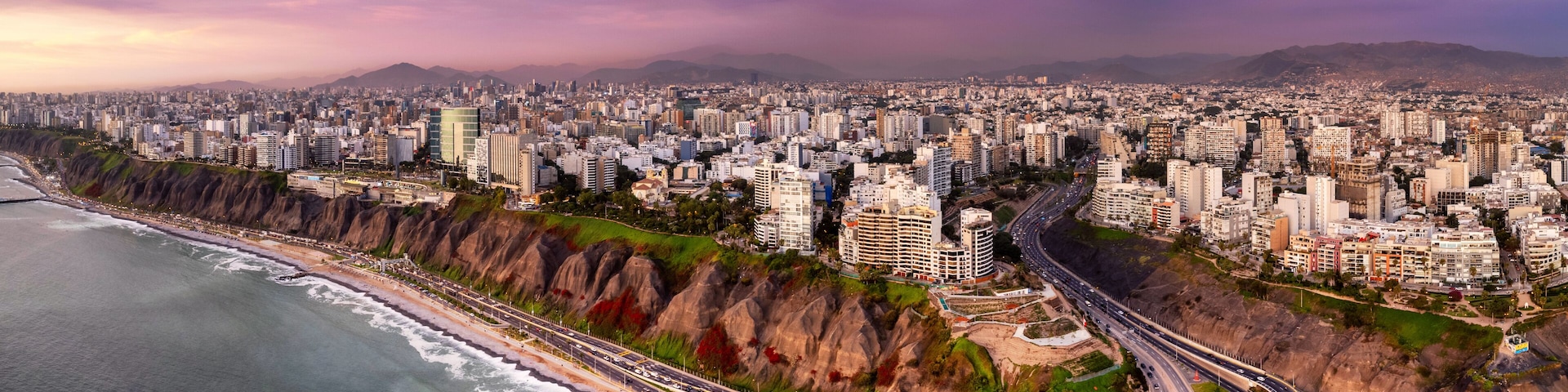 A coastal cityscape with a prominent highway curving along the coastline, dense urban buildings, and a mountain range in the distance.