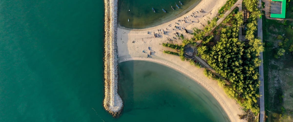 An aerial view of a coastal area with a pier extending into the water, a sandy beach, and a road with buildings.