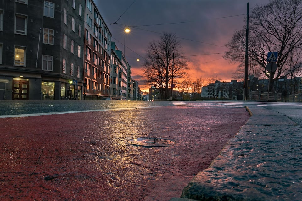 Rua molhada na cidade ao entardecer, com edifícios e pôr do sol.