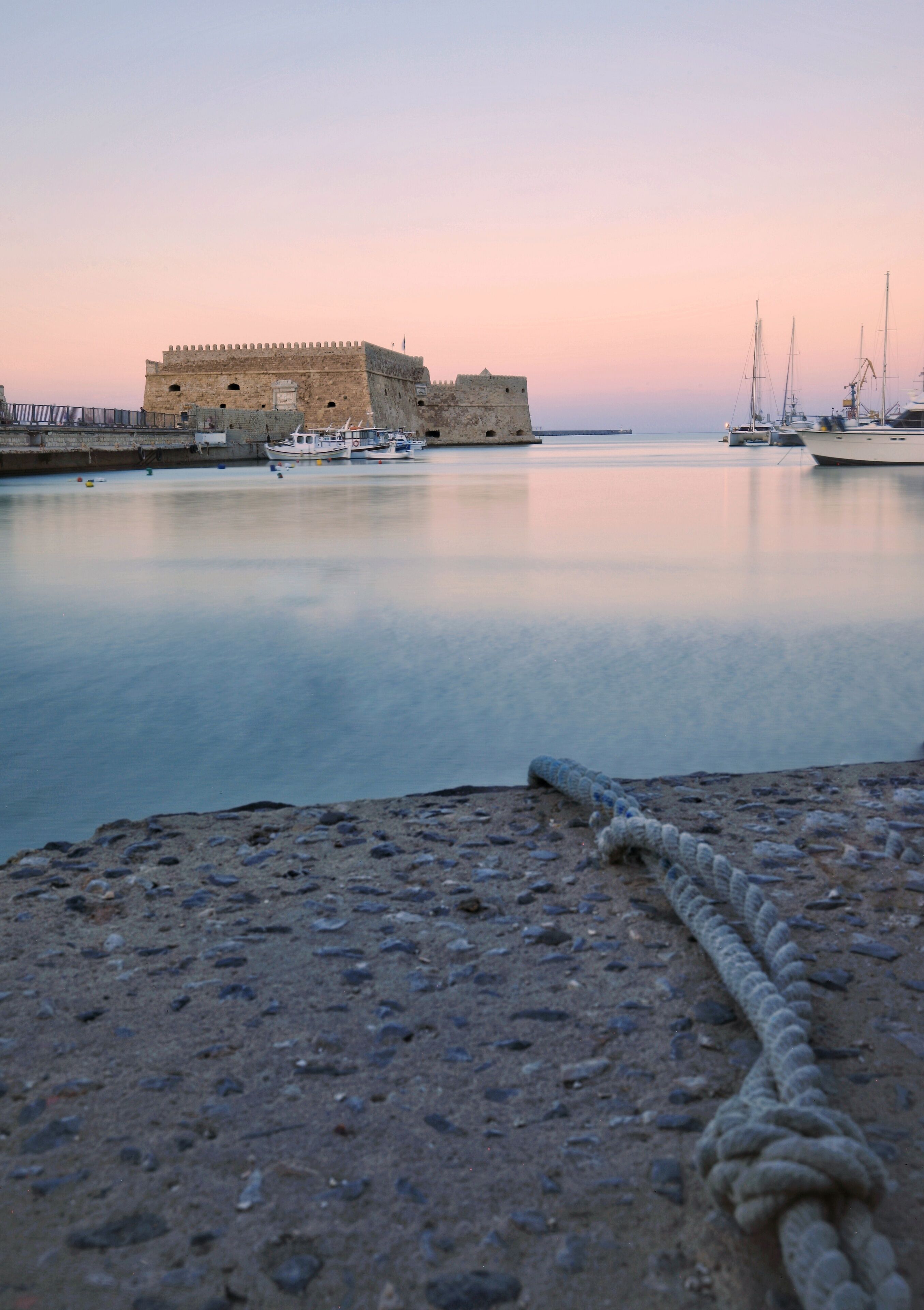 Escena costera con un fuerte histórico, agua tranquila y barcos fondeados.