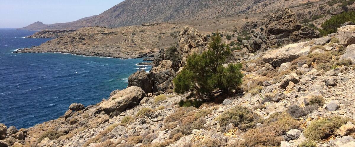 A rocky coastal landscape with a clear blue sea and distant mountains.