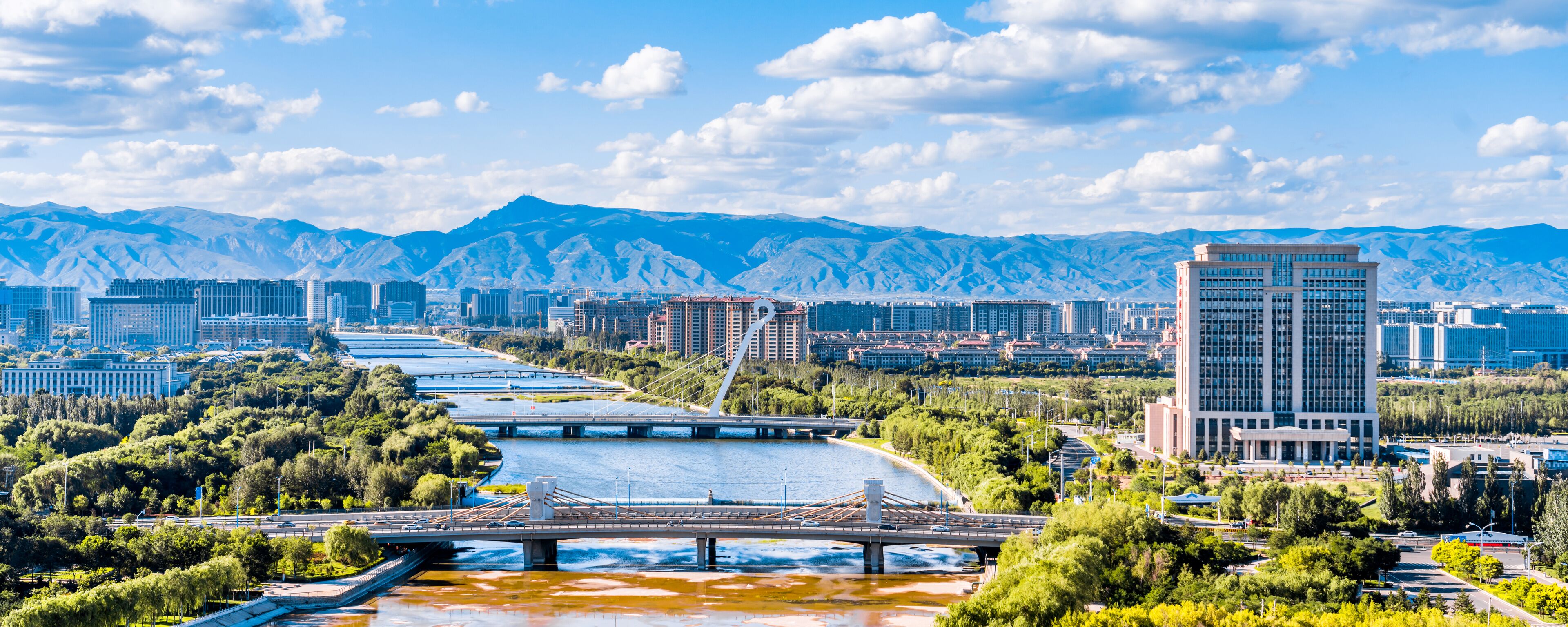 A river with a bridge, a cityscape with buildings, and mountains in the background.