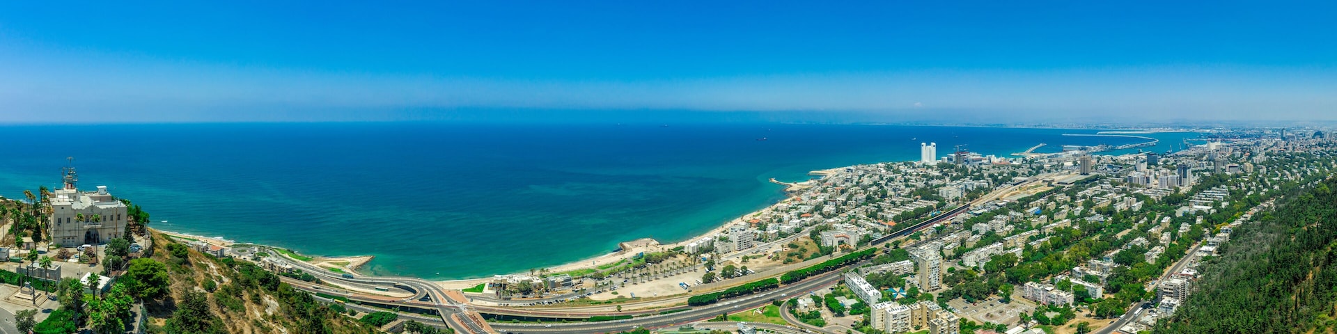 Paisagem costeira com um edifício imponente numa colina, praia e uma área urbana densa com ruas e vegetação.