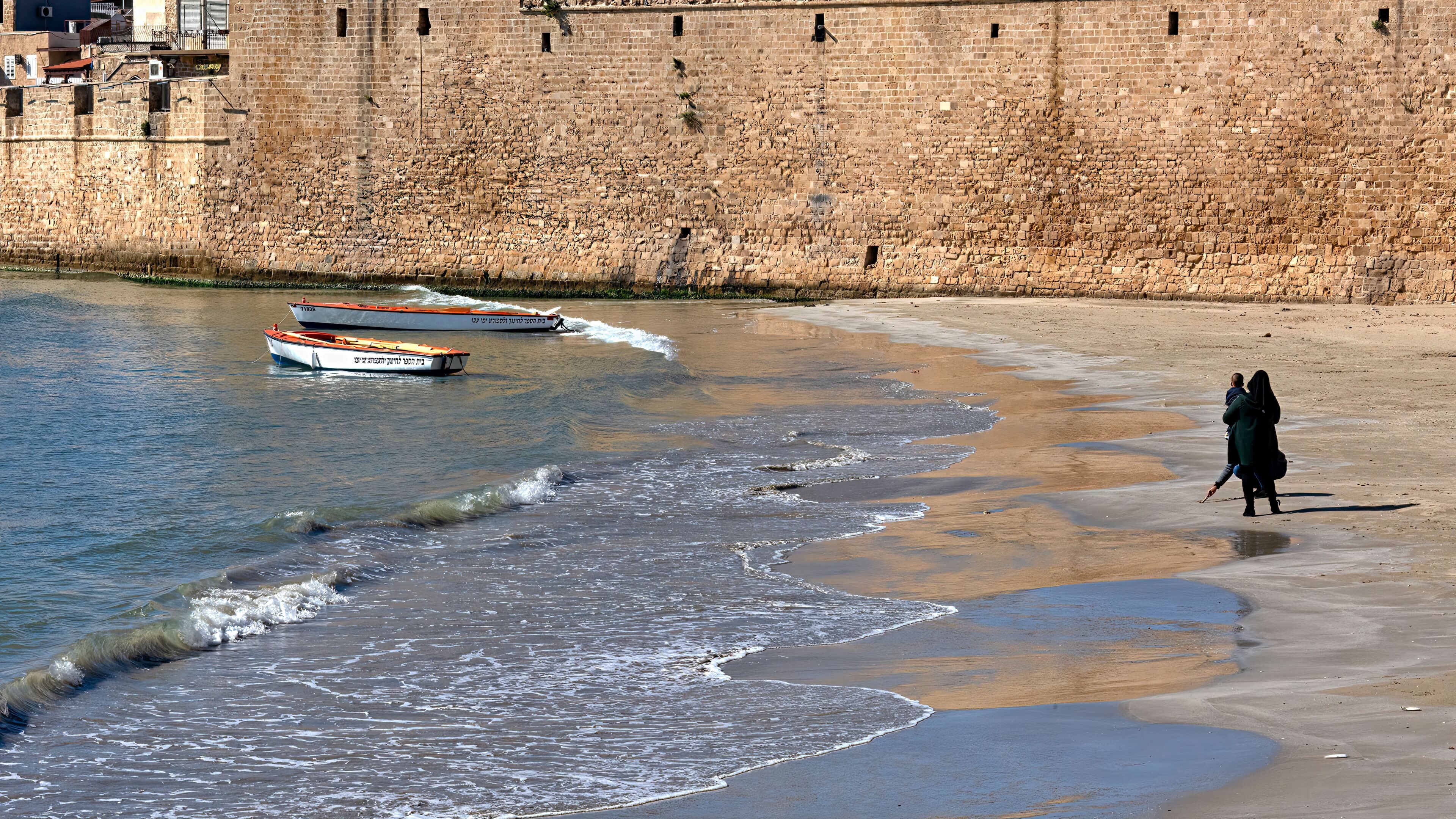 Una playa con olas, barcos y un muro de piedra.