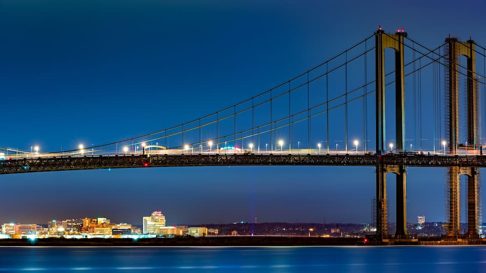 A nighttime view of a large suspension bridge with illuminated cables and towers, spanning over a body of water with a cityscape in the background.