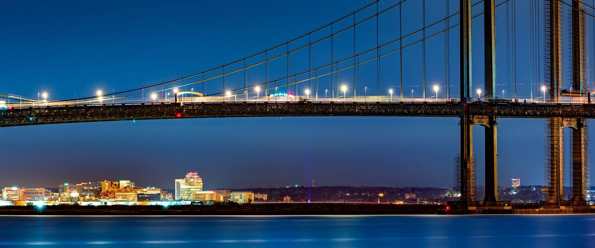 A nighttime view of a large suspension bridge with illuminated cables and towers, spanning over a body of water with a cityscape in the background.