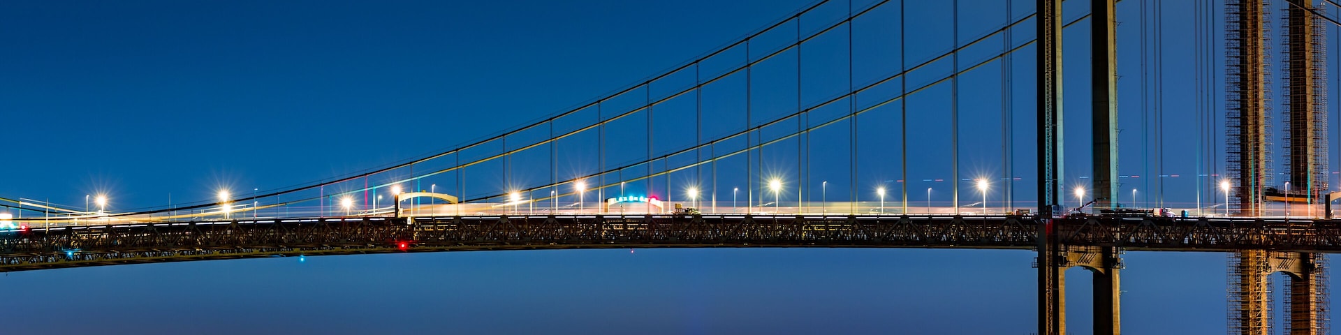 A nighttime view of a large suspension bridge with illuminated cables and towers, spanning over a body of water with a cityscape in the background.