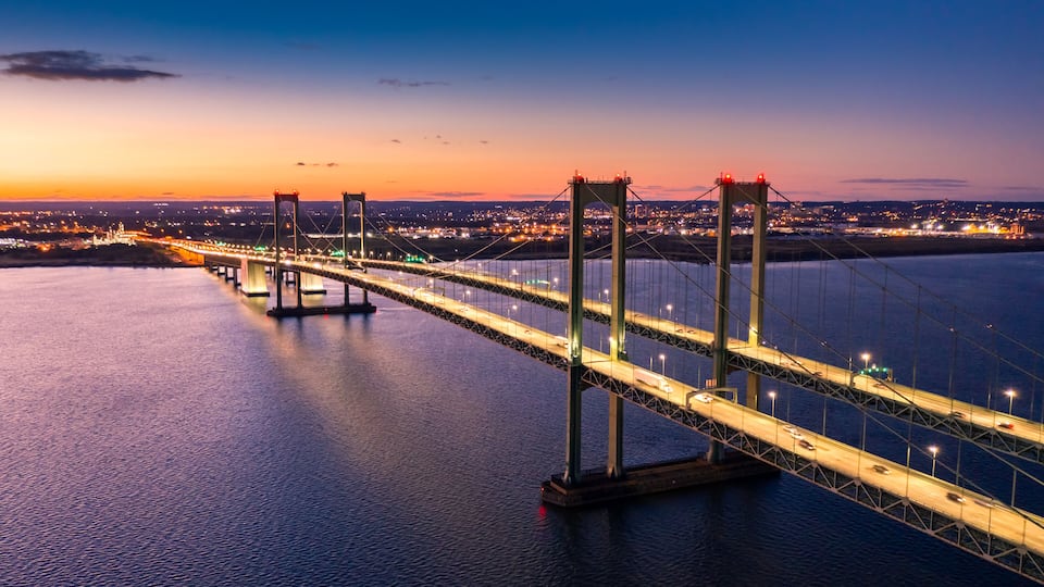 A large suspension bridge at dusk with illuminated roadways and a calm body of water below.