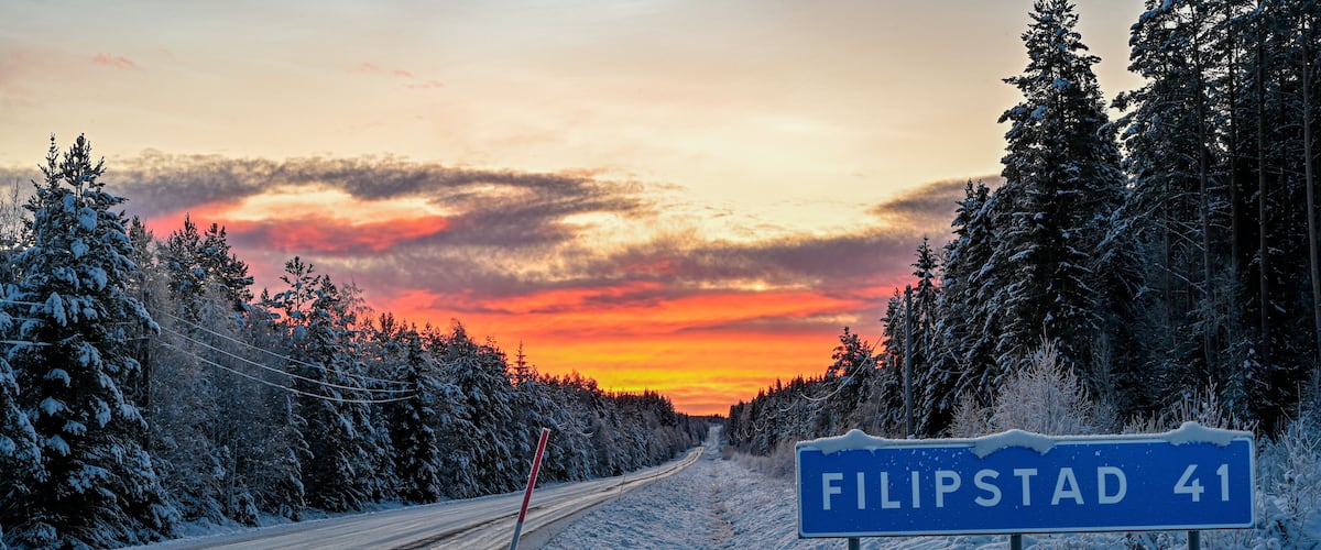 A snow-covered road with a sign for 'Filipstad 41' and a sunset in the background.