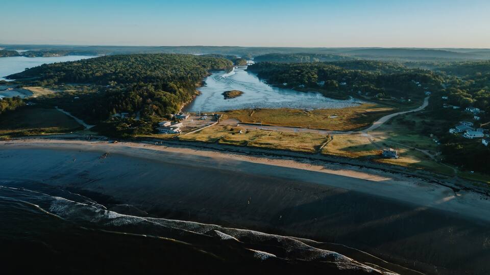 A coastal landscape with a river, forested hills, and a beach.