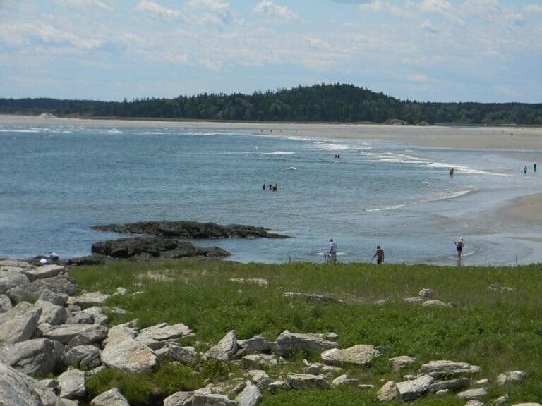 A rocky shoreline with waves crashing onto the beach, a forested hill in the background, and people walking along the shore.