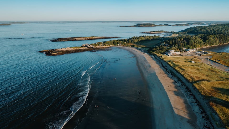 An aerial view of a coastal area with a sandy beach, a road, and a small settlement with buildings.